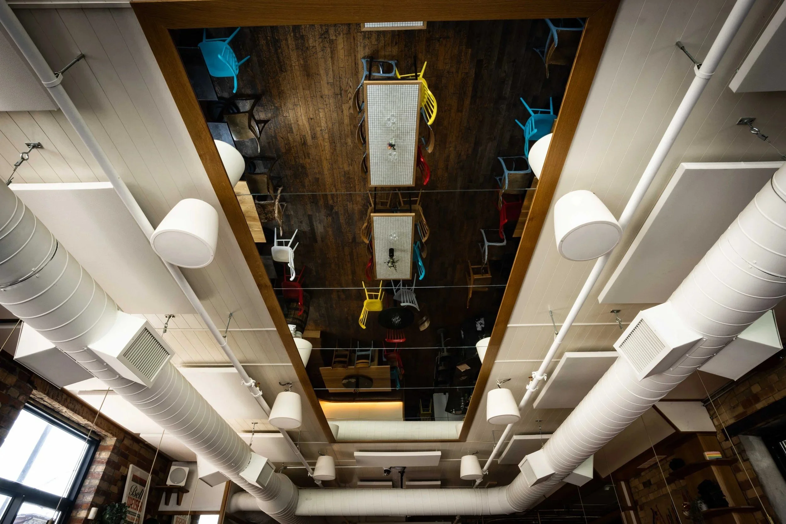 Mirrored ceiling panel reflecting tiled dining tables and colorful chairs below, framed by exposed ductwork, suspended speakers, and modern industrial lighting in a contemporary restaurant interior.