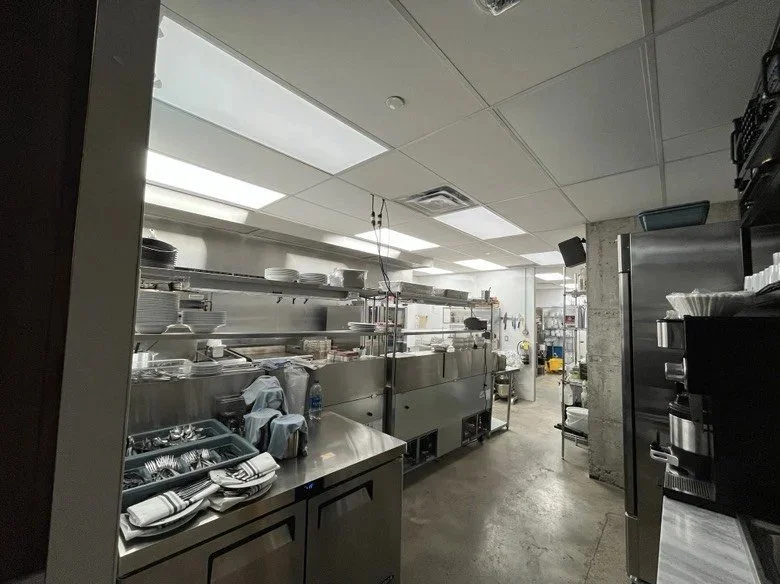 Commercial restaurant kitchen with stainless steel cooking line, prep counters, refrigeration, shelving, dishware, and food service equipment illuminated by bright overhead lighting.