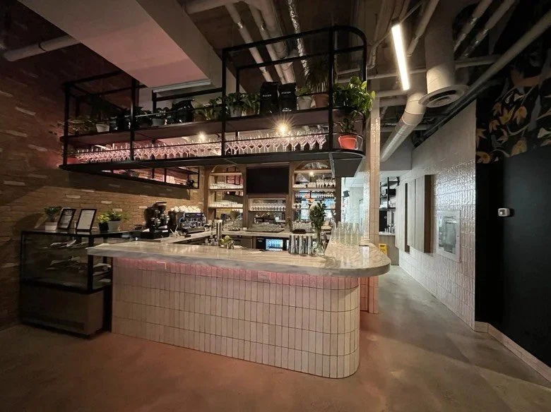 Modern restaurant bar featuring a curved marble countertop, suspended black metal shelving with hanging glassware and plants, exposed brick walls, café equipment, and an open layout connecting the dining area and kitchen.