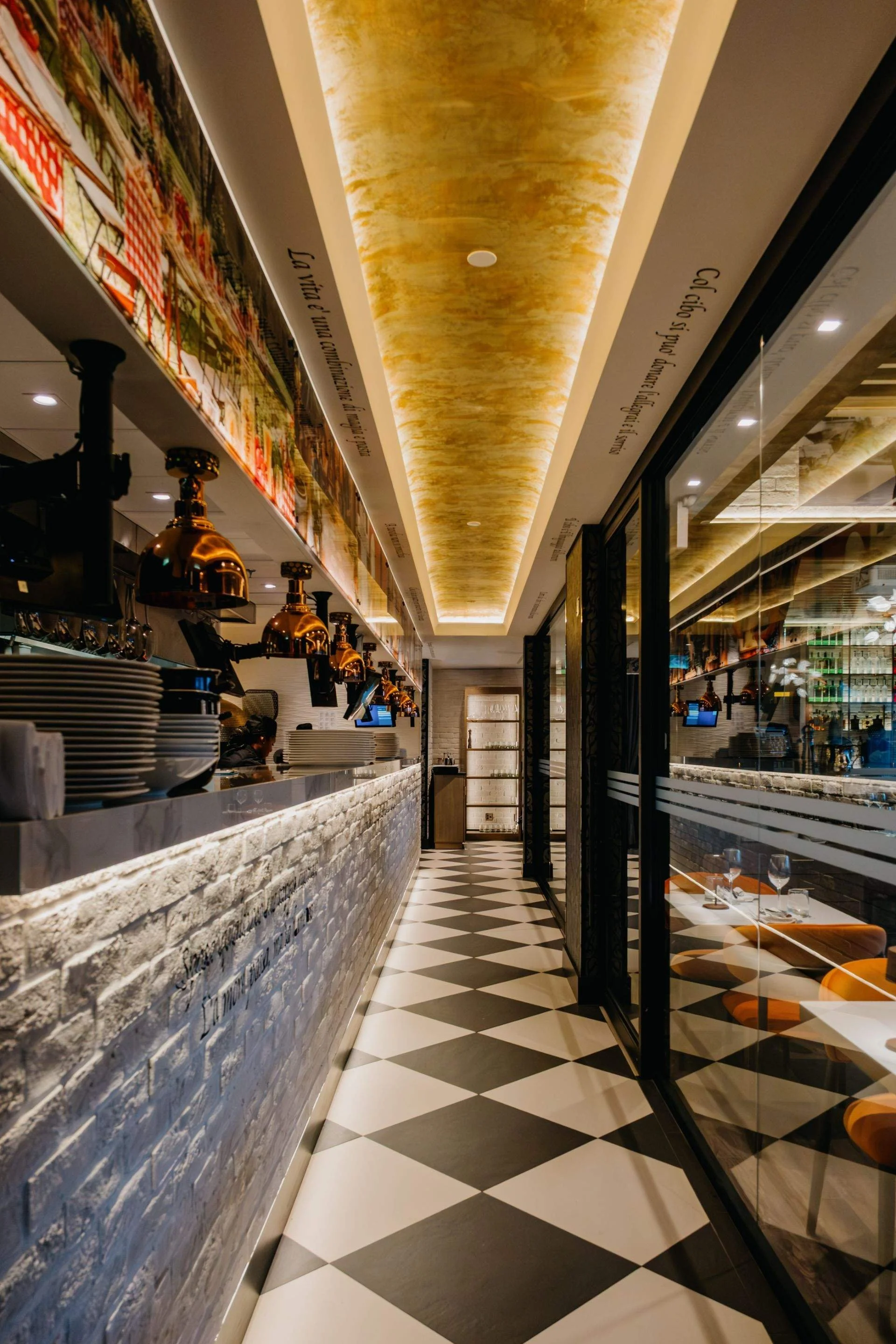 Restaurant kitchen corridor with a black-and-white geometric floor, white brick counter, copper heat lamps, and glass partitions.