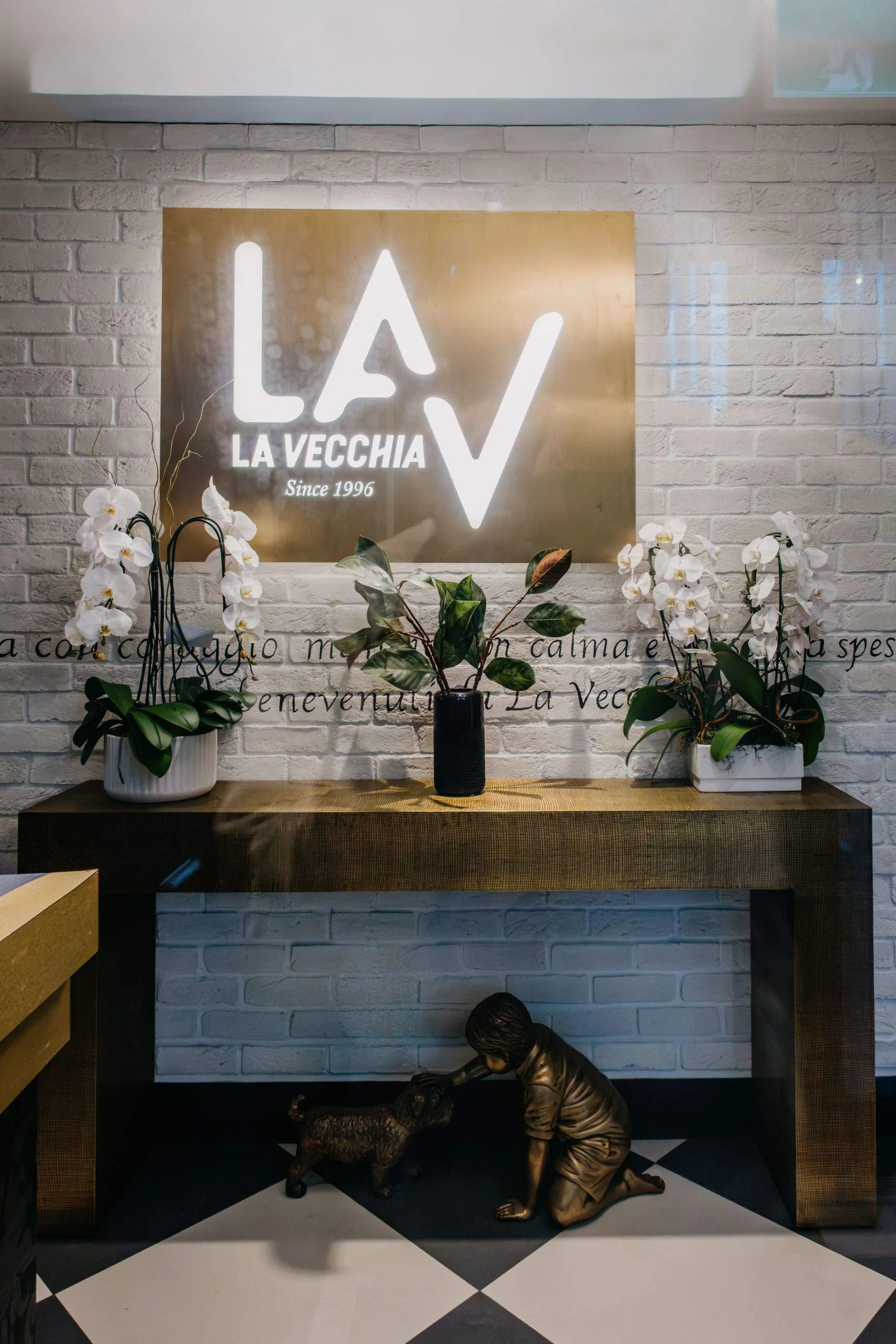 Illuminated La Vecchia “Since 1996” logo on a white brick wall above orchids and a decorative sculpture.