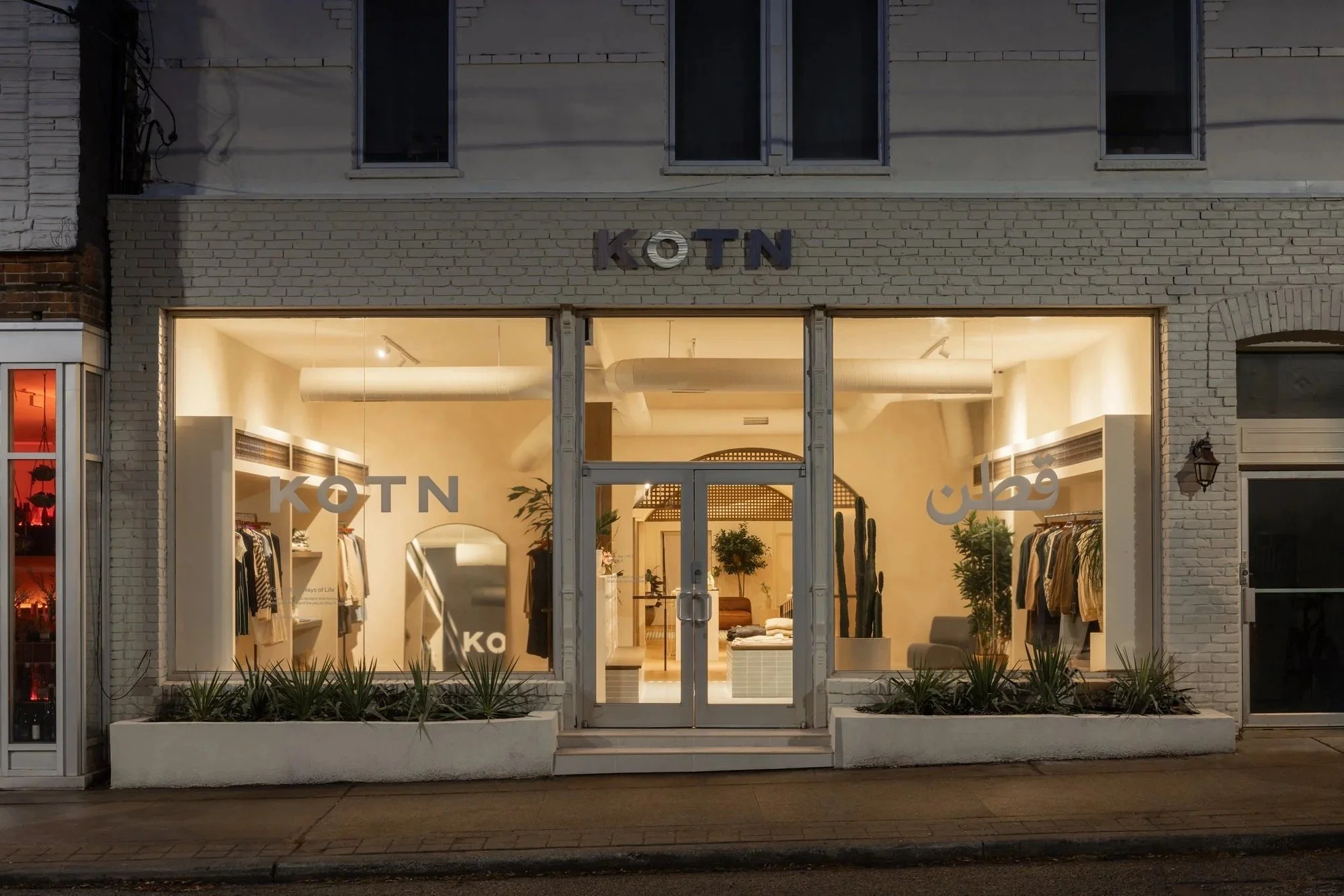 Exterior view of the KOTN clothing store at night, featuring a bright, minimalist storefront with large display windows, clothing racks, indoor plants, illuminated signage, and a modern retail interior visible from the street.