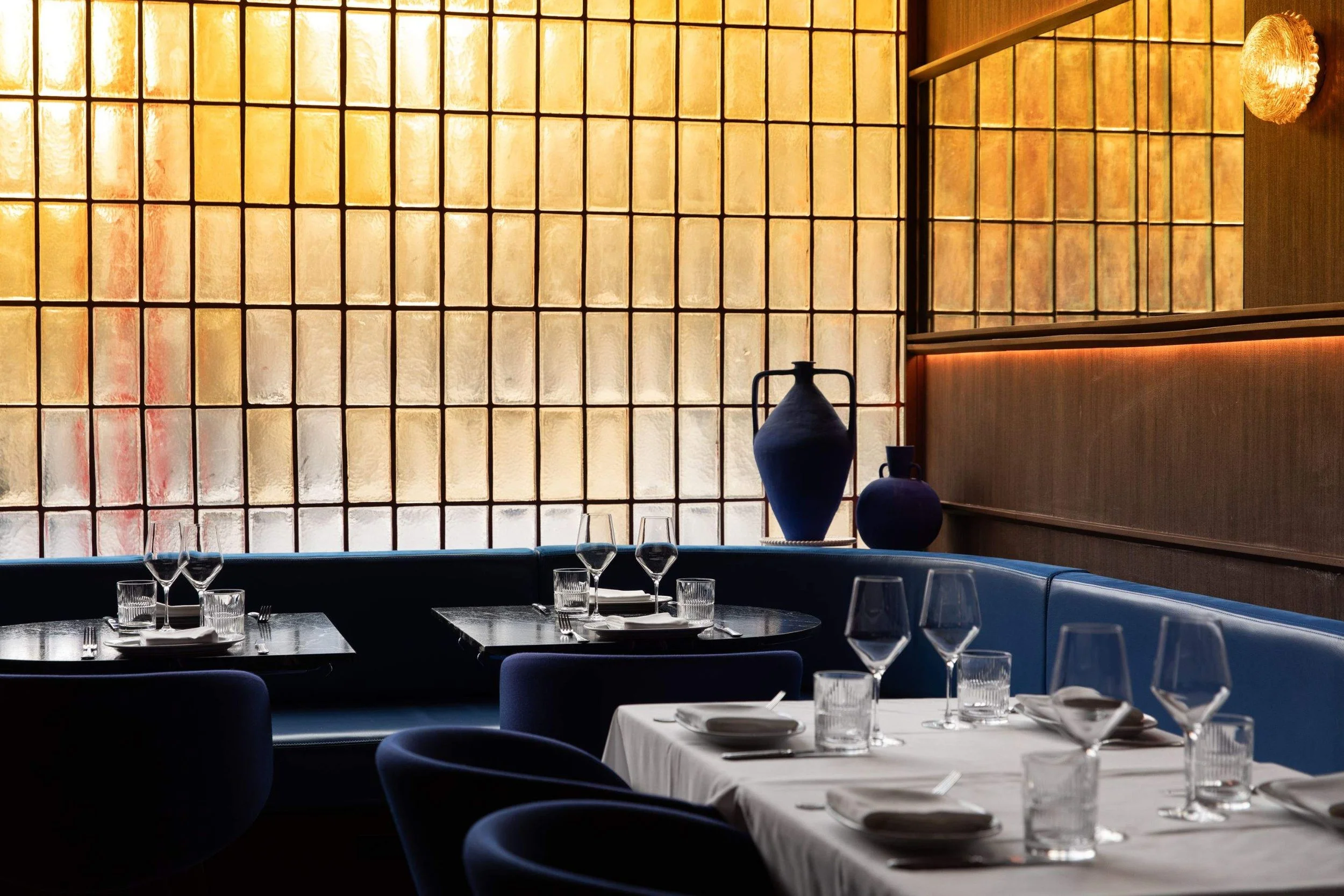 Intimate restaurant dining area with deep blue seating, white tablecloths, decorative blue vases, and a large amber-tinted glass-block window.