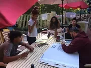 Participants making posters around an outdoor table beneath umbrellas.