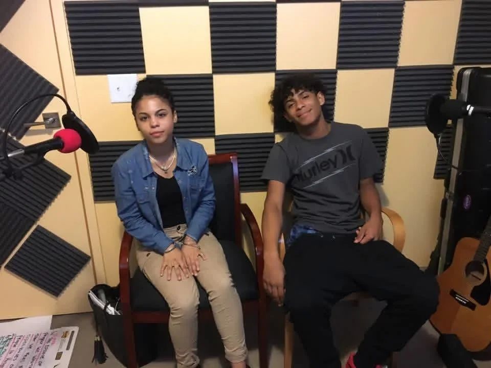 Two participants seated beside microphones in a recording booth.