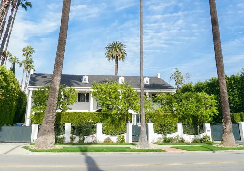 A white two-story home behind a hedge on a Los Angeles street lined with tall palms
