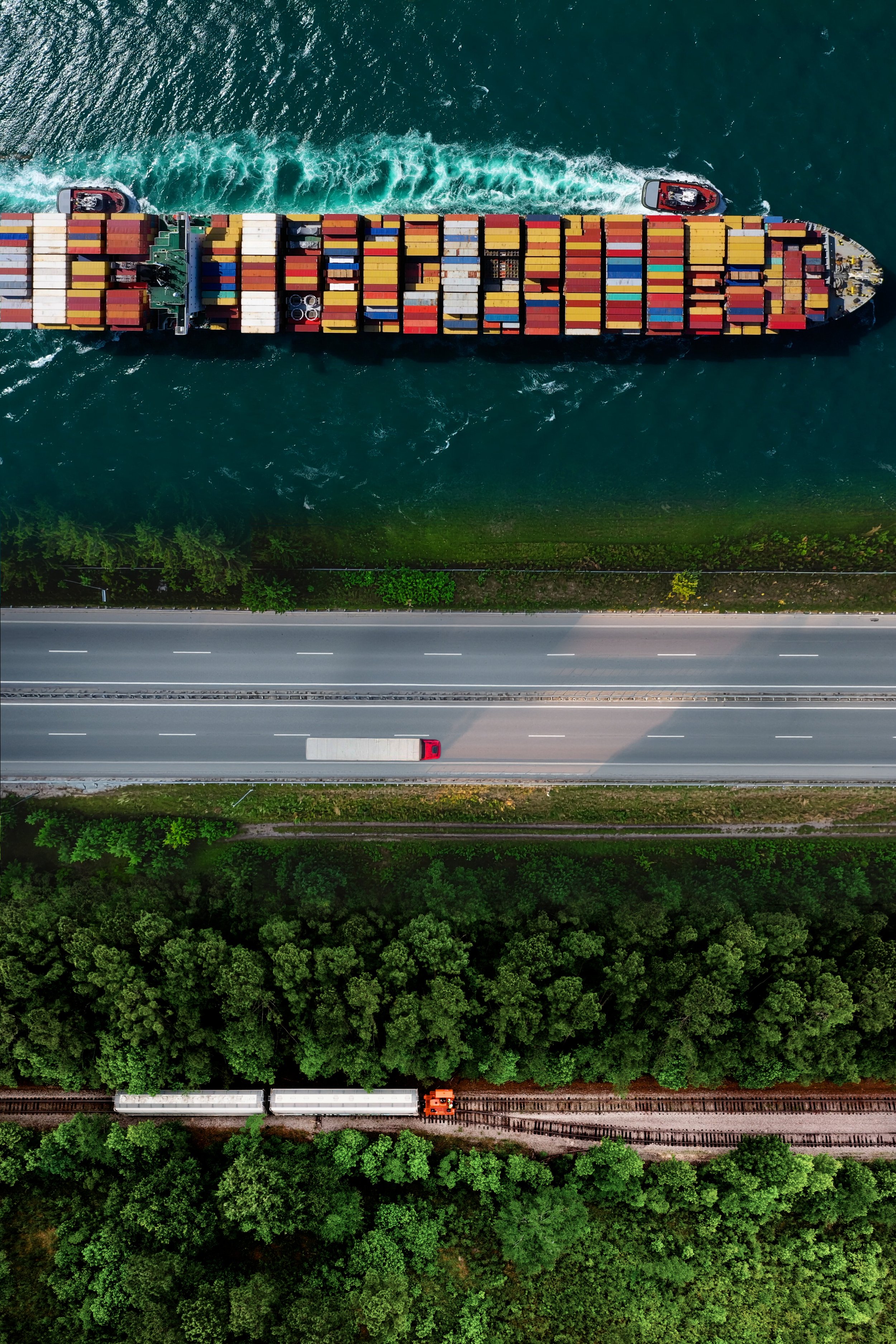 Aerial view of a container ship, highway, and freight train running parallel through green landscape