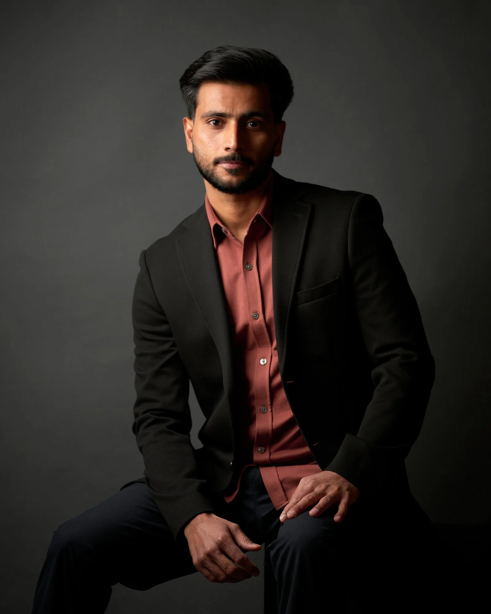Actor in a black blazer and rust shirt seated against dark grey