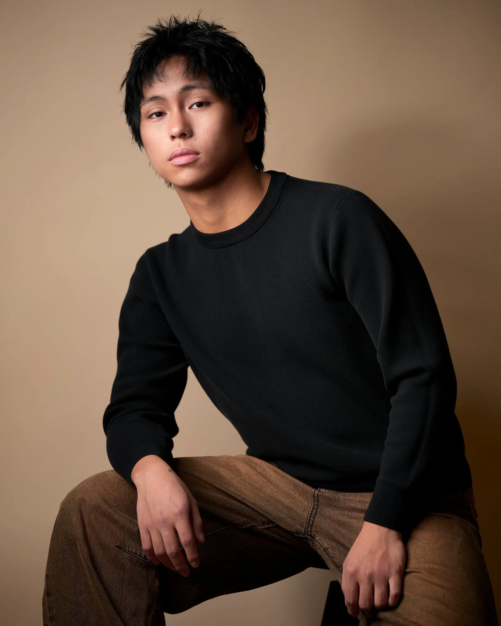 Young actor in a black sweater and brown trousers seated against tan