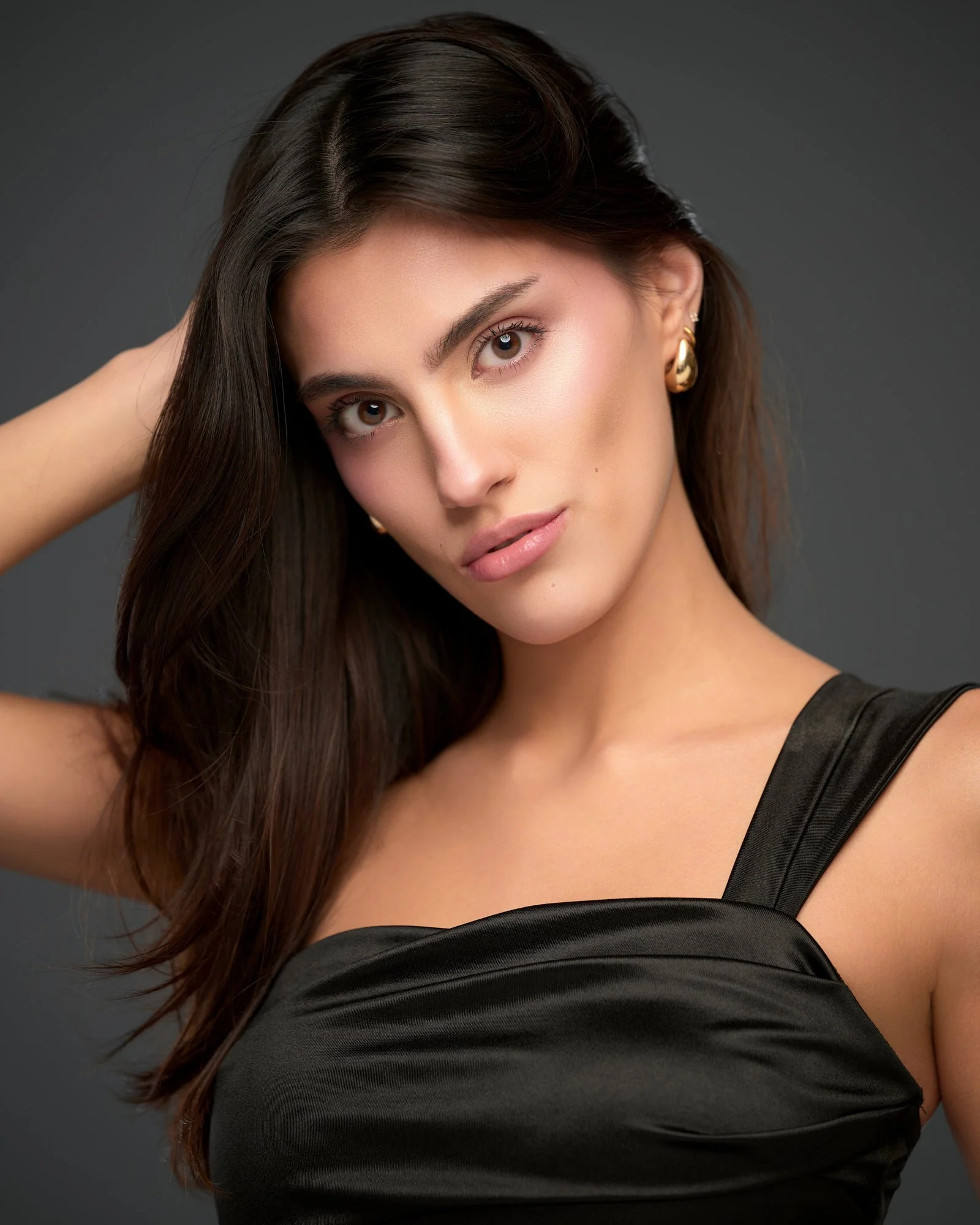 Model headshot in a black satin top and gold earrings on dark grey