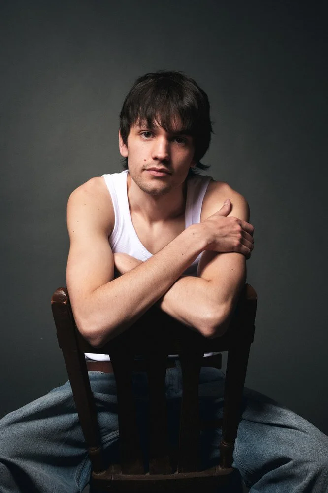 Male model in a white tank top seated on a chair against a dark backdrop