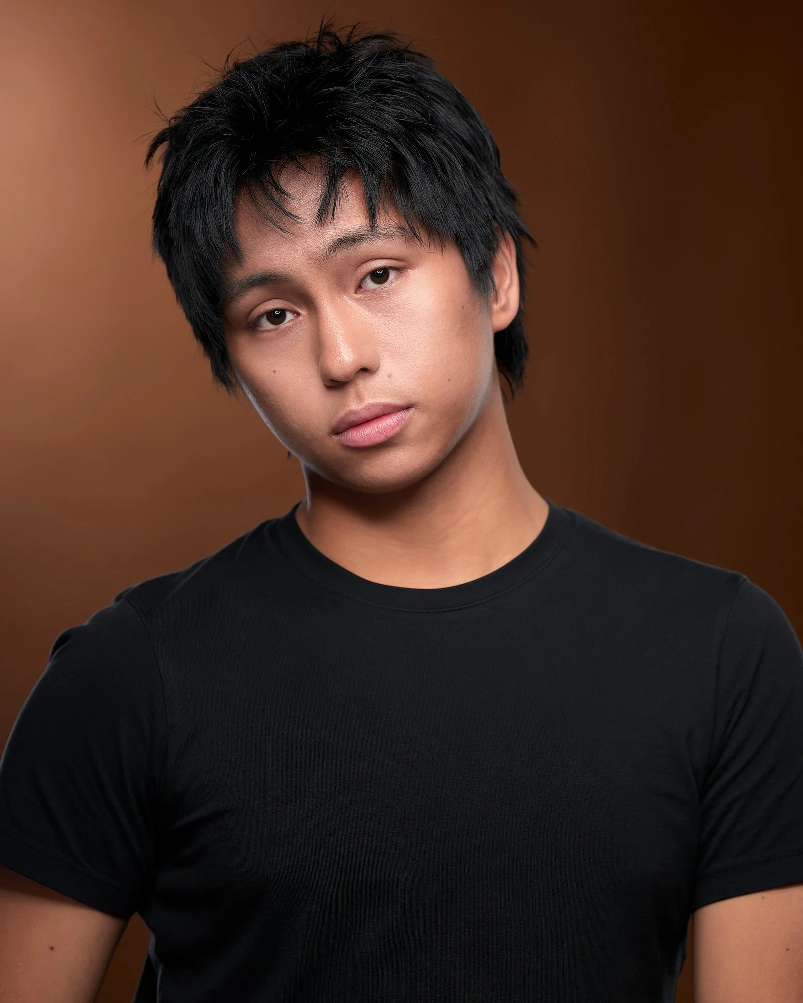 Young actor in a black tee with a head tilt on a warm brown backdrop