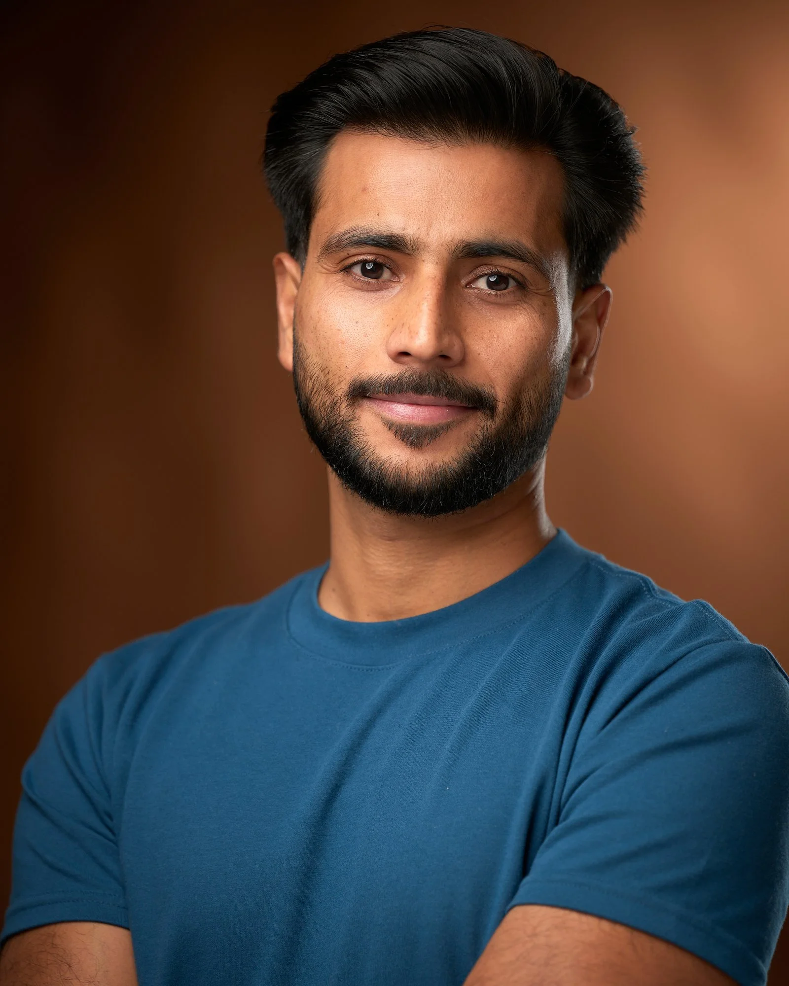 Actor in a blue tee with arms crossed against a warm brown backdrop