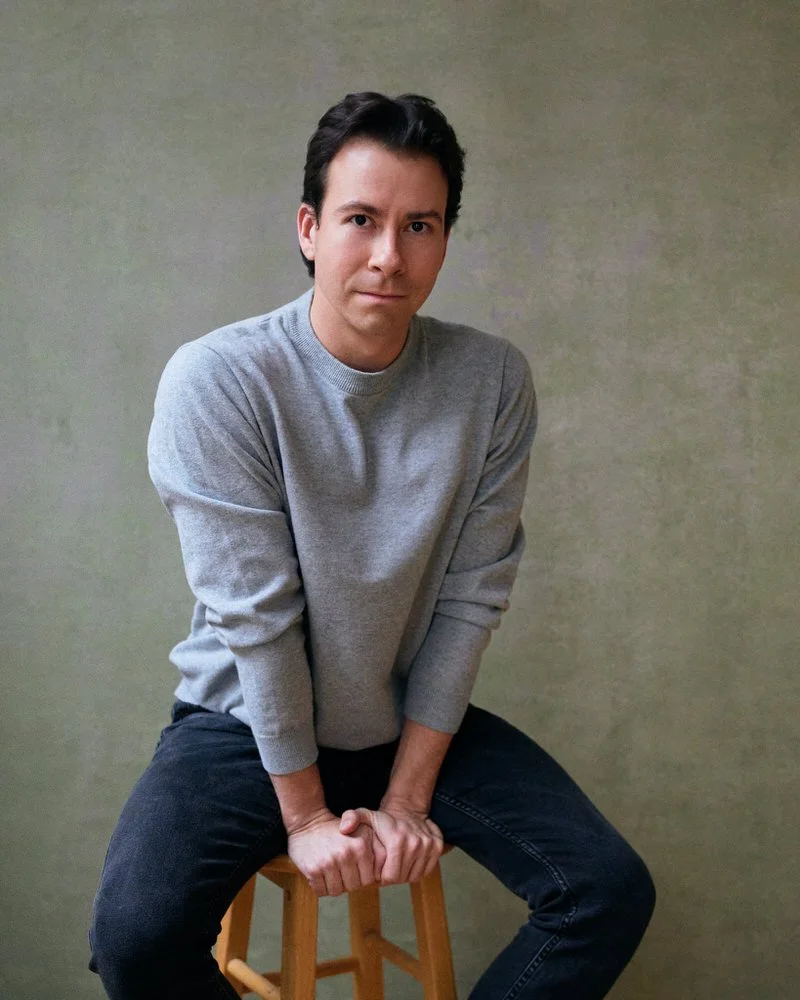 Actor in a grey sweater seated against a textured backdrop