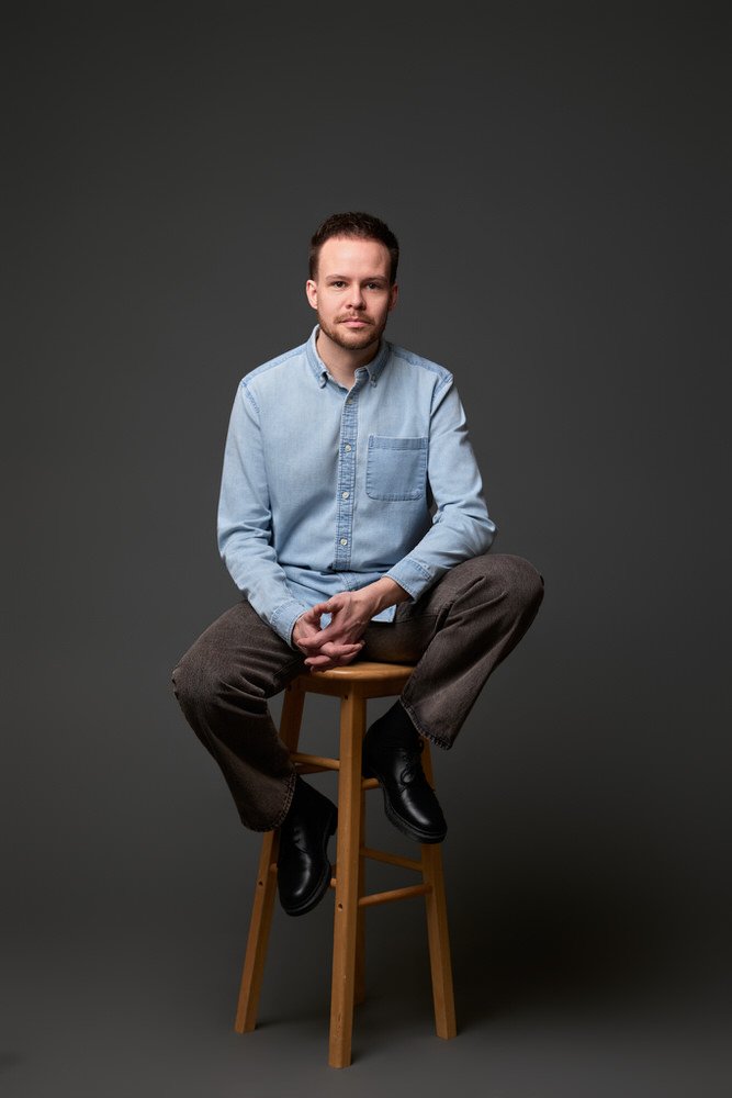 Full-length image of an actor in a denim shirt seated on grey