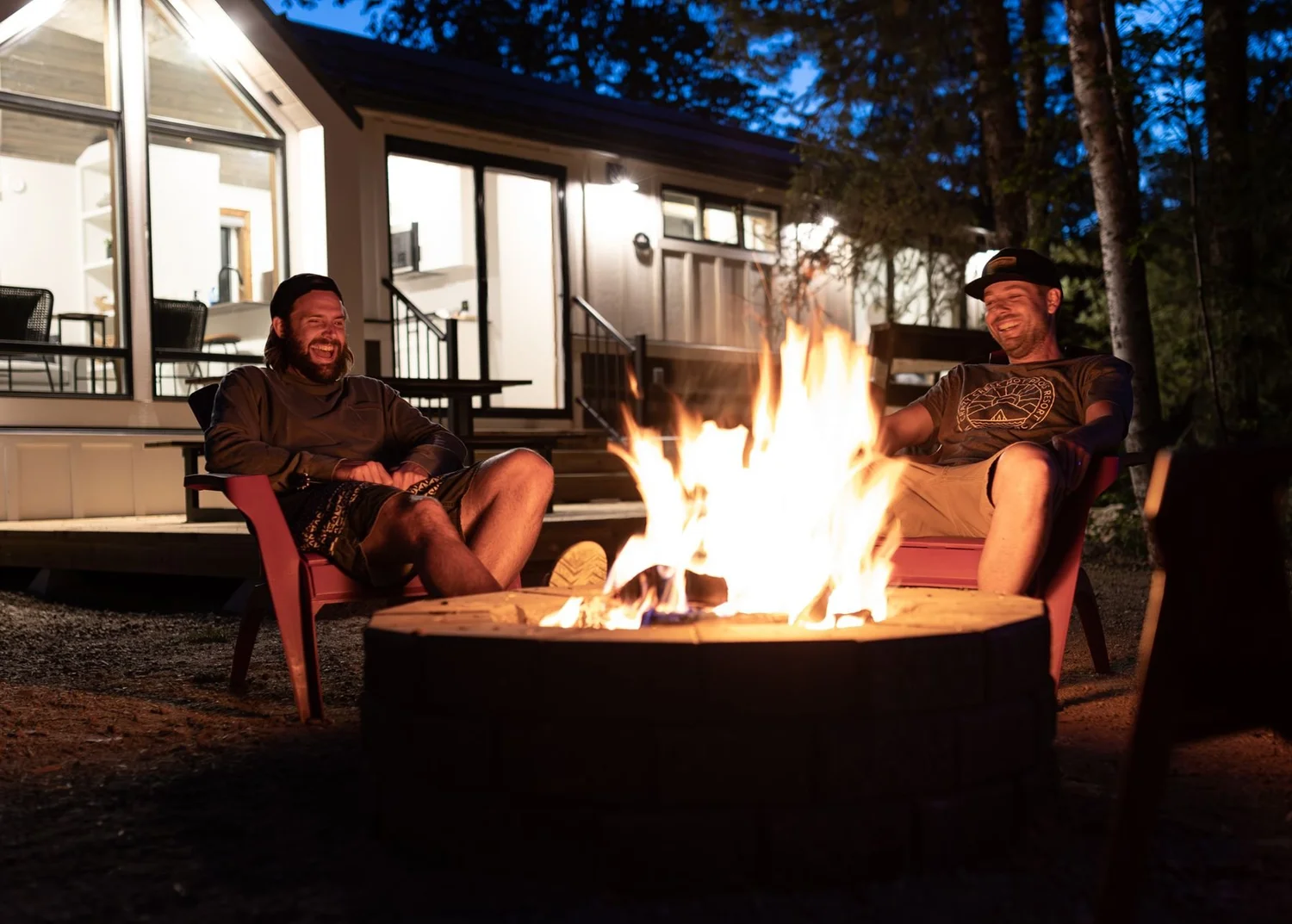 Two friends laughing in red Adirondack chairs beside a roaring fire pit at night, a lit Crazy Creek cabin behind them