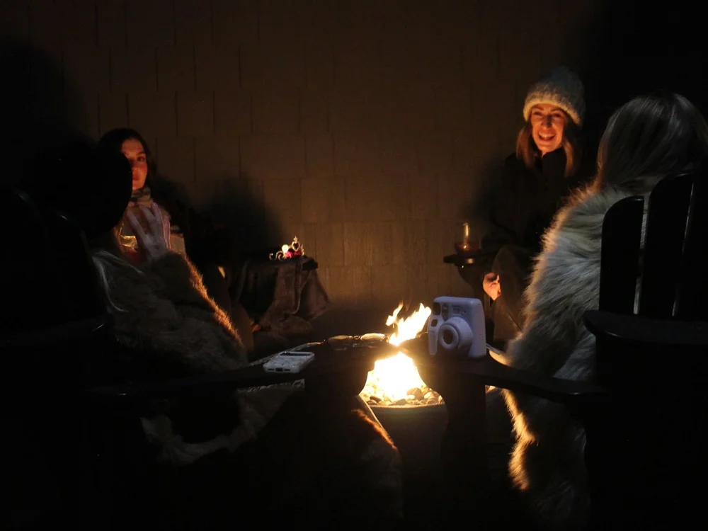 Friends in toques and warm coats laughing around a small fire at night