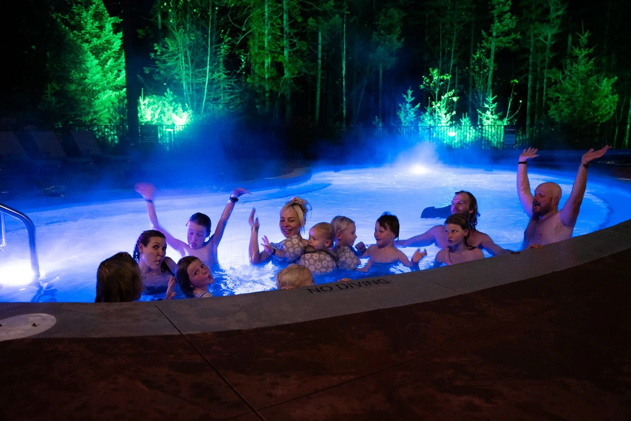 A big group of parents and kids cheering with their arms up in the hot pool at night, steam rising under the lit trees