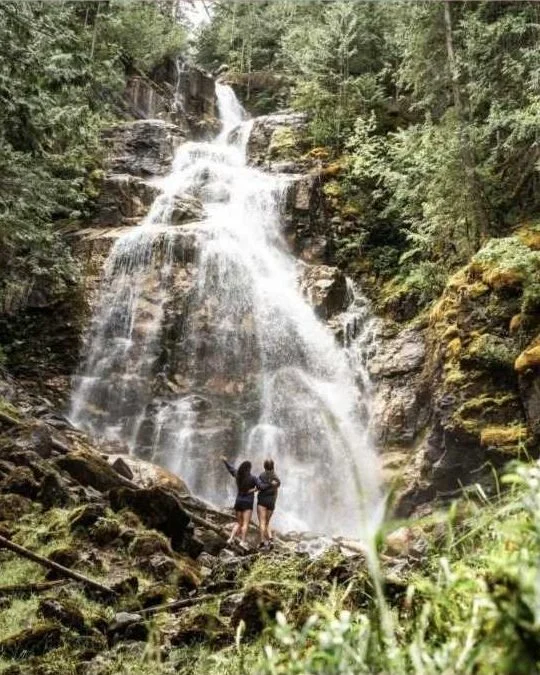 Two people with arms raised at the base of Kay Falls as it pours over mossy rock in summer