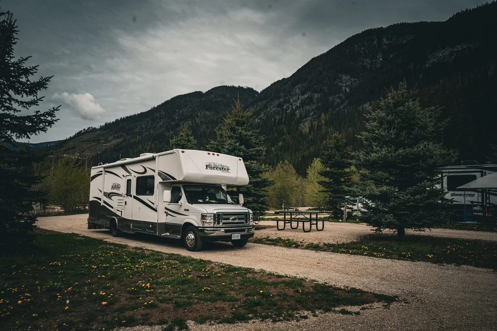Class C motorhome parked on a long gravel pull-through site, with forested mountains behind