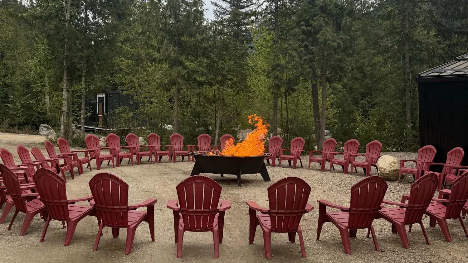 A ring of red Adirondack chairs facing a fire pit with flames rising, tall cedars and firs behind