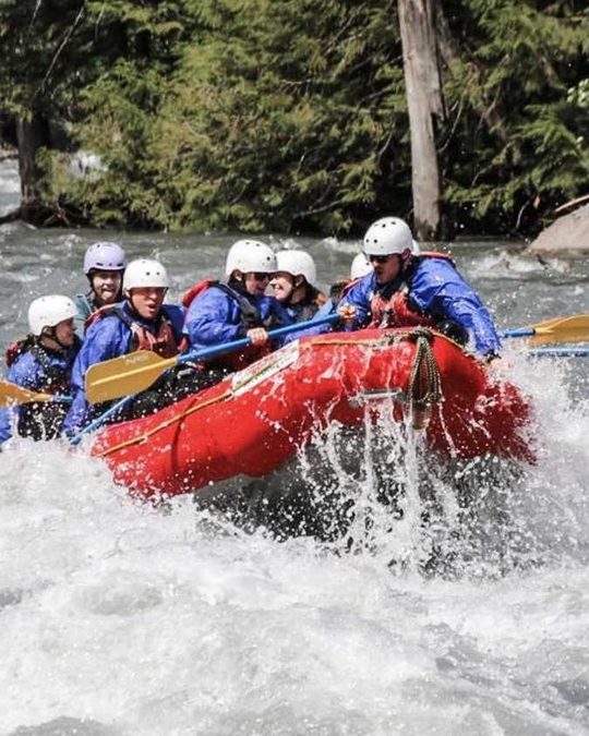 A red raft full of paddlers in helmets crashing through whitewater on a forested river