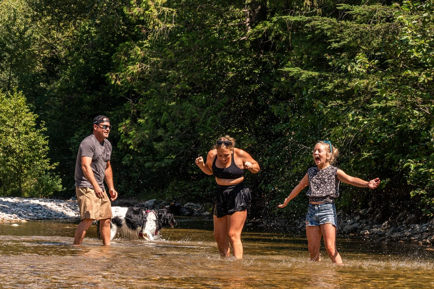 A family laughing and splashing in the shallow, clear water of the Eagle River while their dog plays nearby, with forest behind