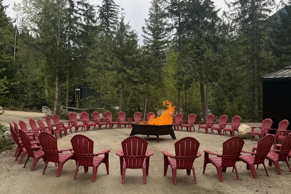 A ring of red Adirondack chairs around a lit fire pit in the forest at Crazy Creek