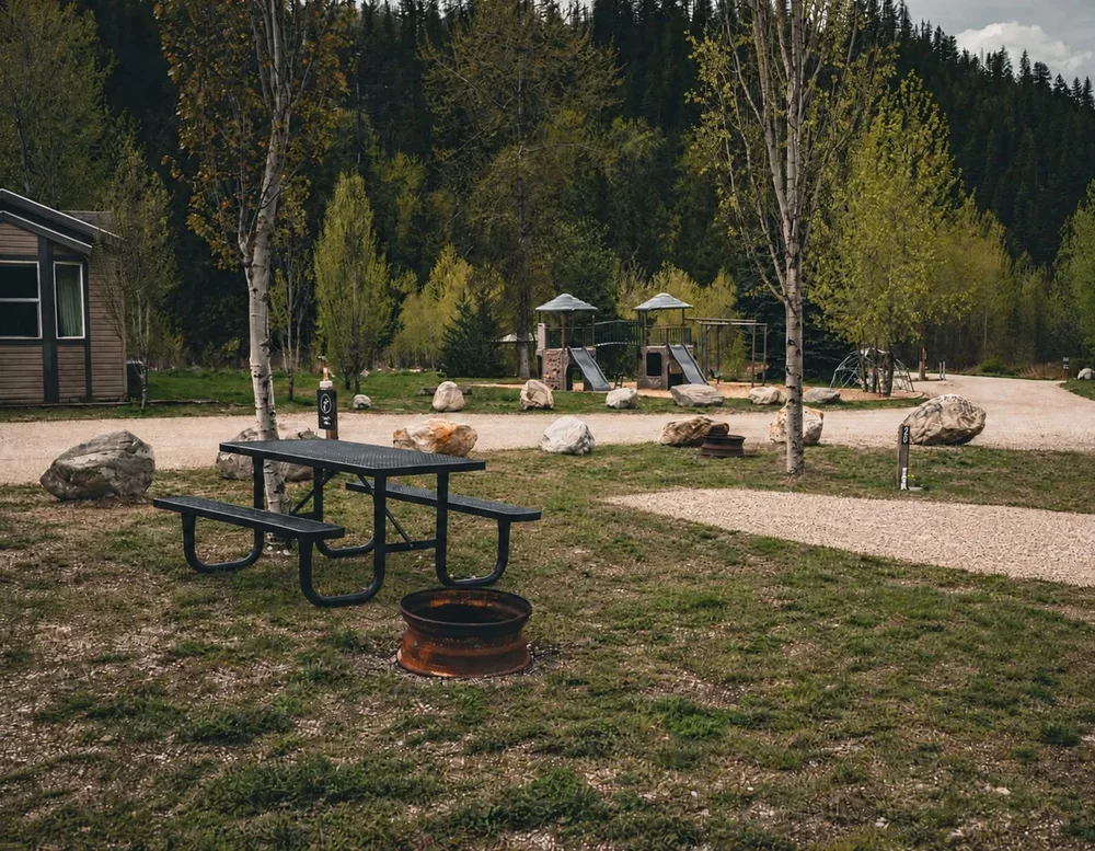 Grassy campsite with a picnic table and fire ring, a cabin to one side and the playground just across the road