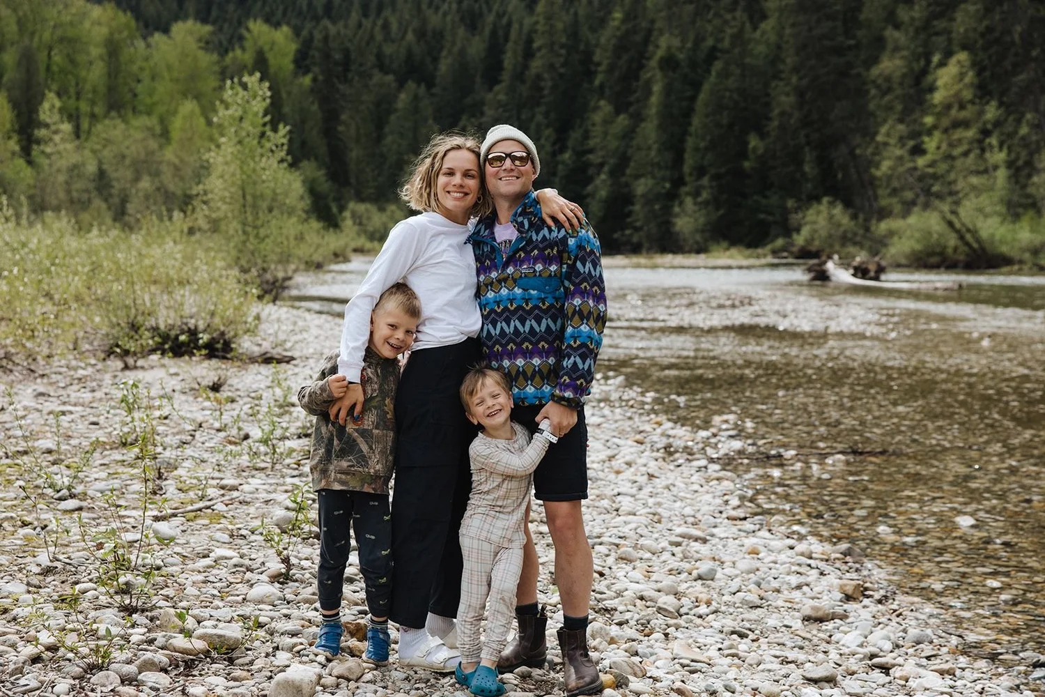 A family of four hugging on a gravel bar beside the Eagle River, with forest and mountains behind