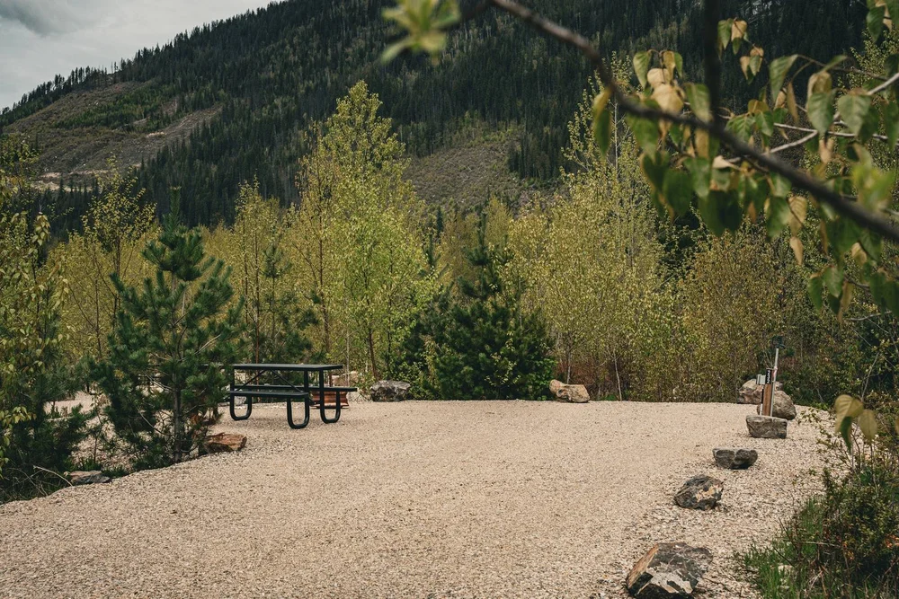 Spacious gravel RV site with a picnic table, edged by boulders and young trees below a forested mountain