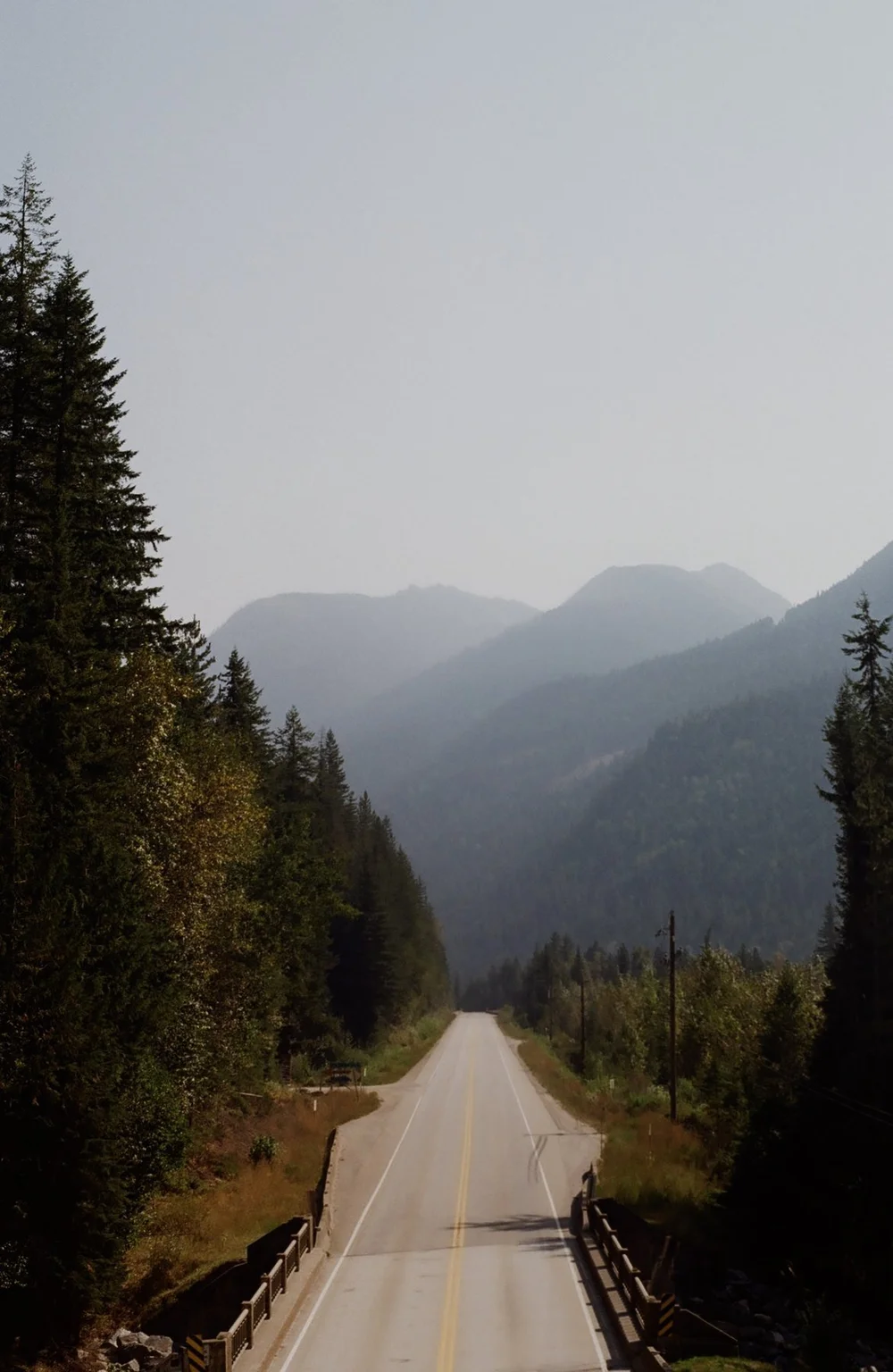 A quiet two-lane road running between tall evergreens toward hazy, forested mountains near Crazy Creek