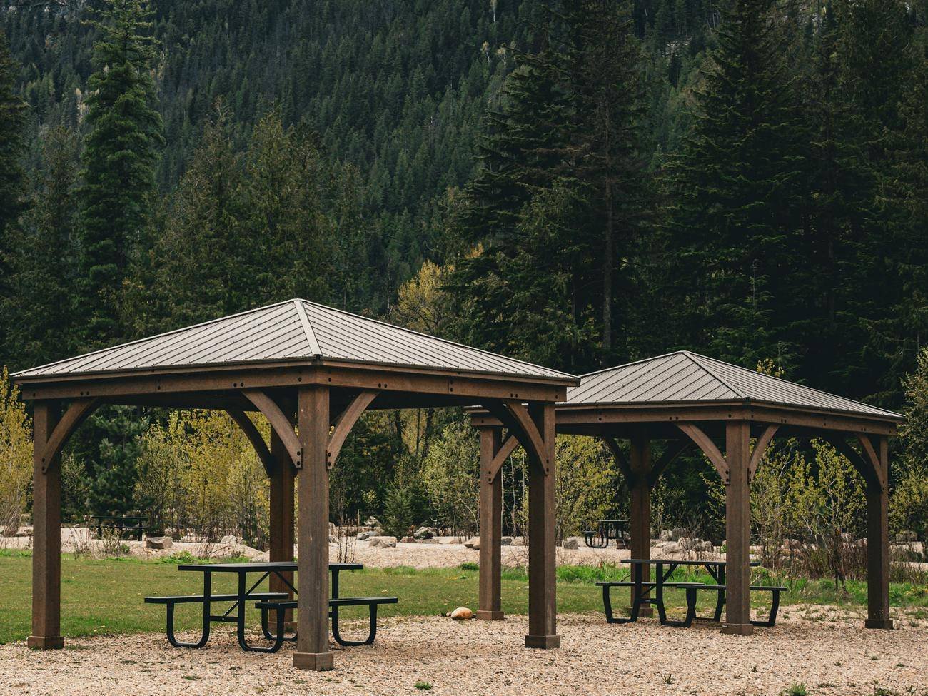 Two wooden cabanas with picnic tables at the edge of the grass field, with the forest behind