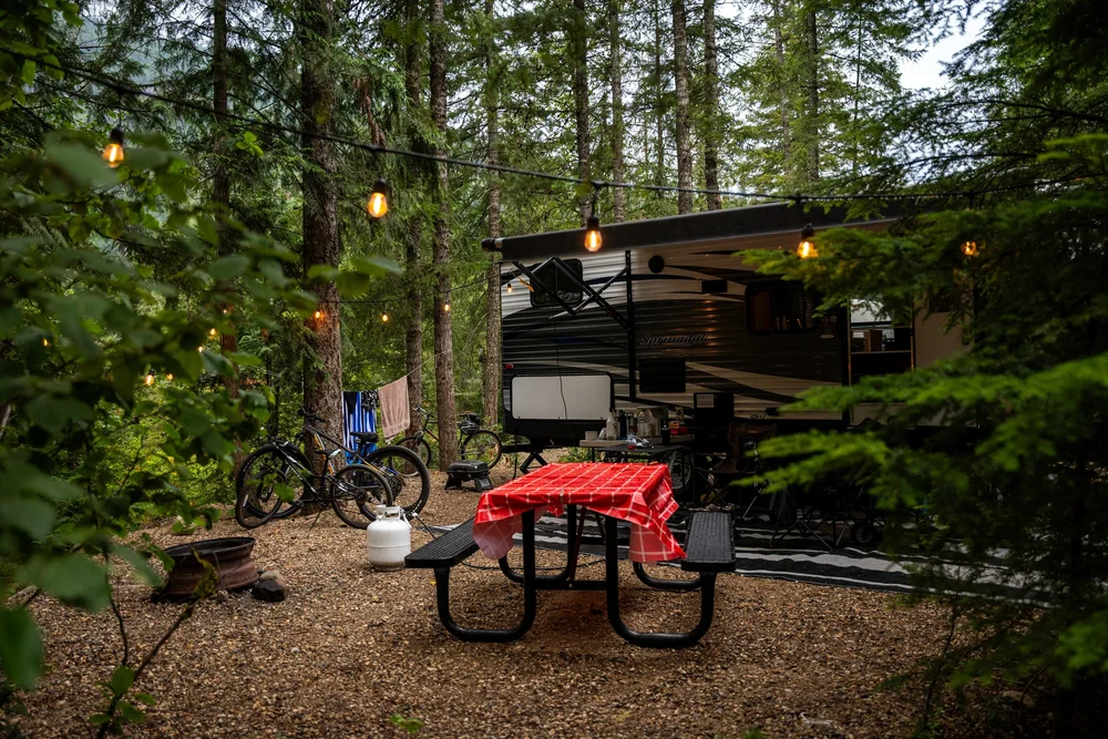 A travel trailer with its awning out at a forest campsite, string lights overhead and a red checked cloth on the picnic table