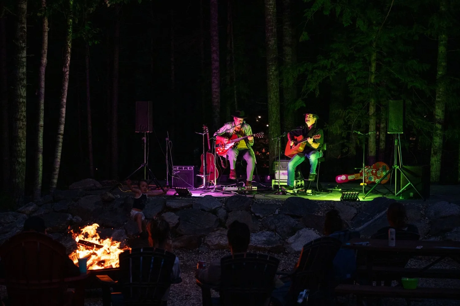 Two musicians playing on the lit outdoor stage among tall trees at night, a campfire glowing in front of the audience