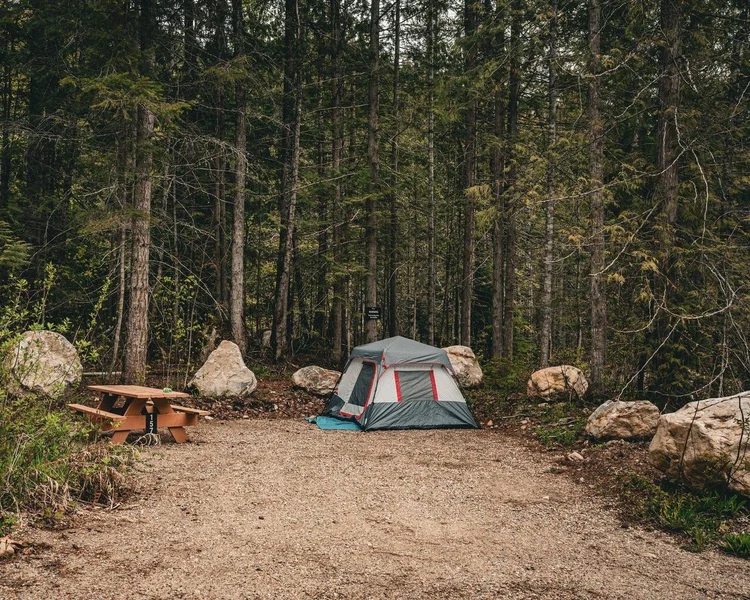 A tent and picnic table on a gravel campsite ringed by boulders and tall forest