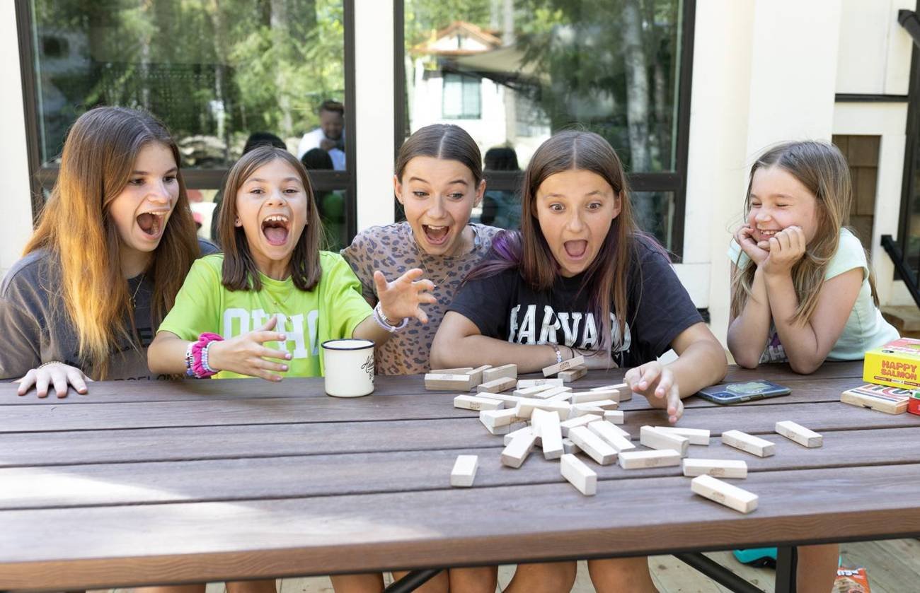 Cousins laughing over a stacking-block game at a picnic table on a cabin deck
