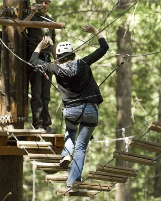 A climber in a helmet crossing a rope bridge between cedar trunks at SkyTrek