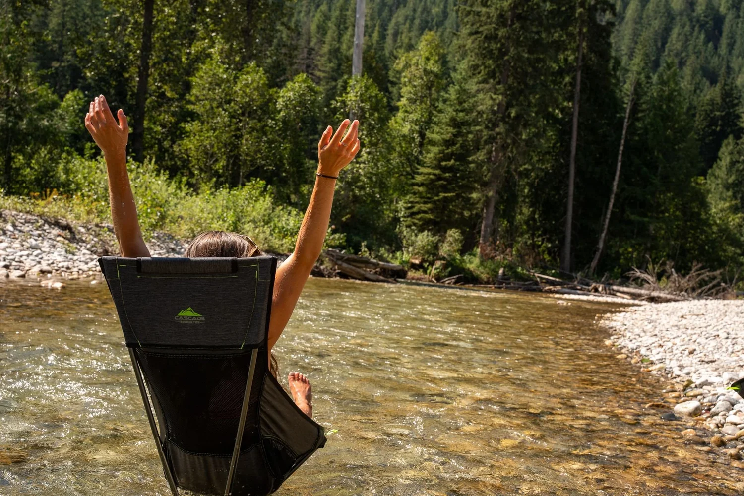 A guest with both arms raised, relaxing in a camp chair in the clear, shallow Eagle River beside a gravel bar, forest behind