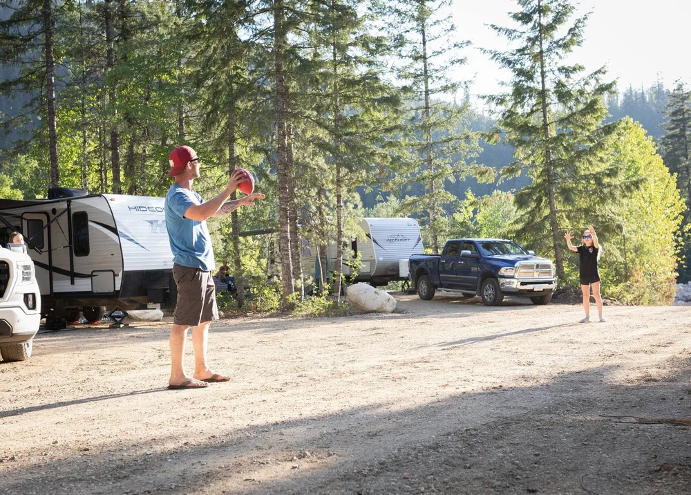 Two campers tossing a football at a shaded forest campsite, with travel trailers and a truck parked among tall trees