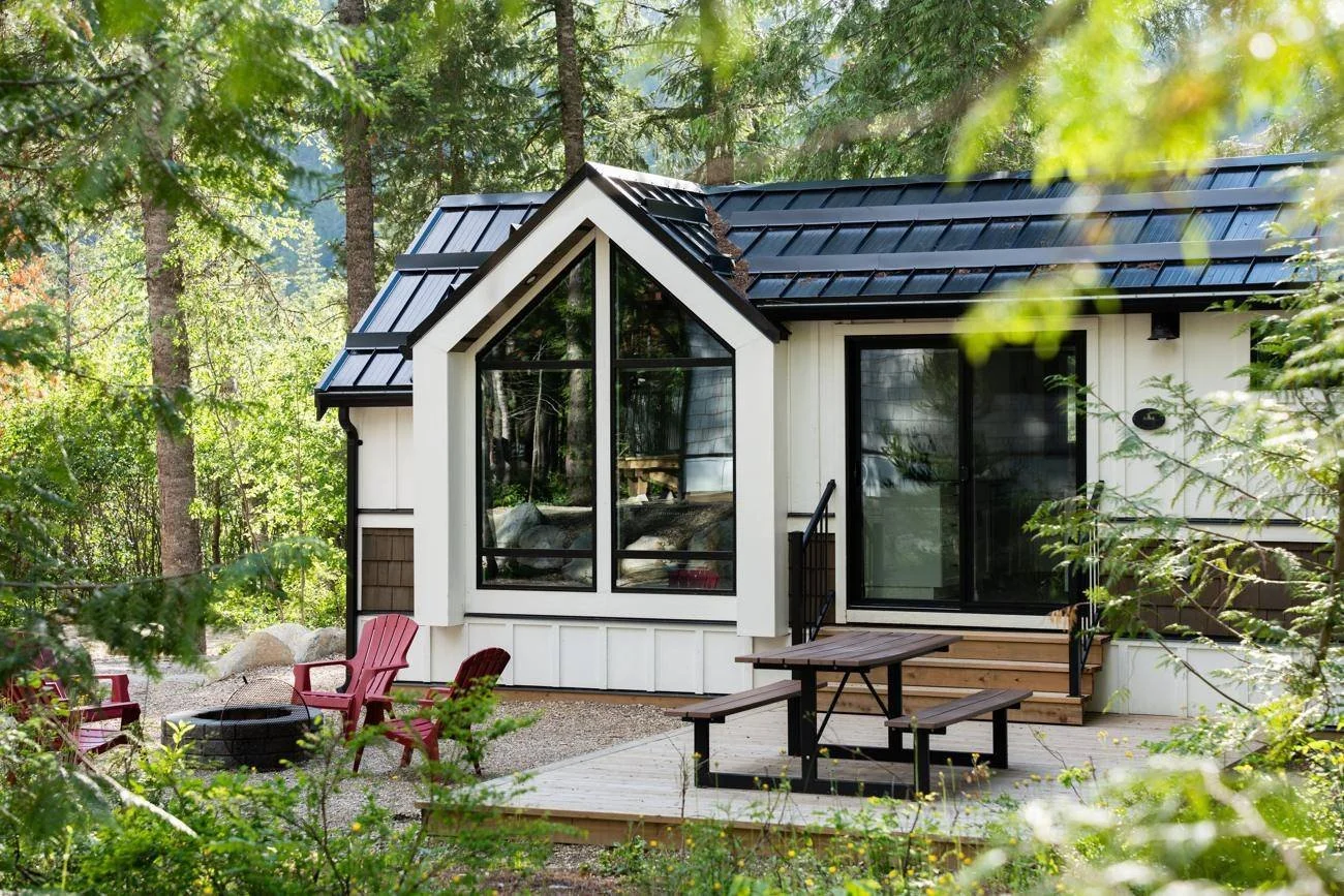 The Apex cabin among the trees, with its tall gabled window wall, red Adirondack chairs around the fire pit and a picnic table on the deck