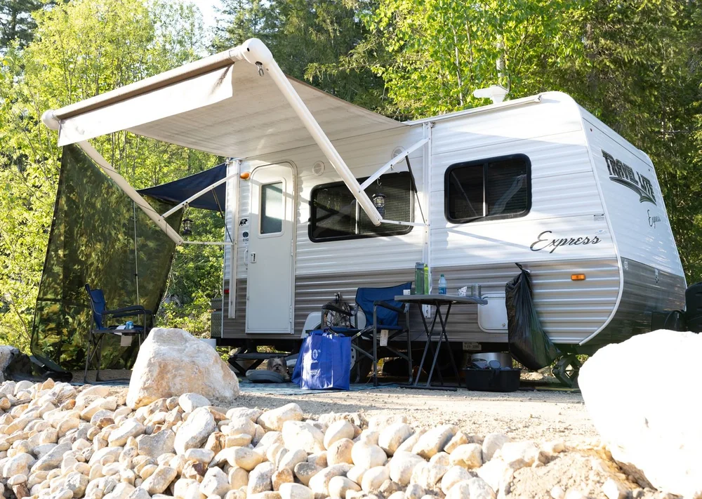 Small travel trailer with its awning out at a forest campsite, a camp chair and cooler beside it