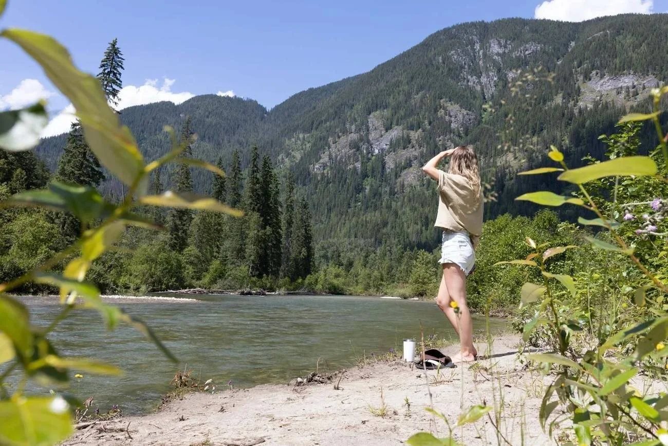A guest on the sandy river beach at Crazy Creek, with forested mountains behind