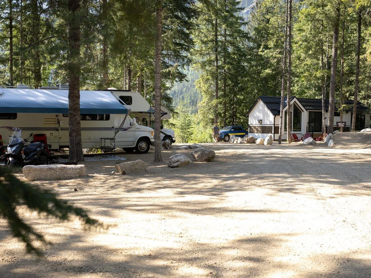 A motorhome with its awning out at a treed RV site, with forest cabins just down the lane