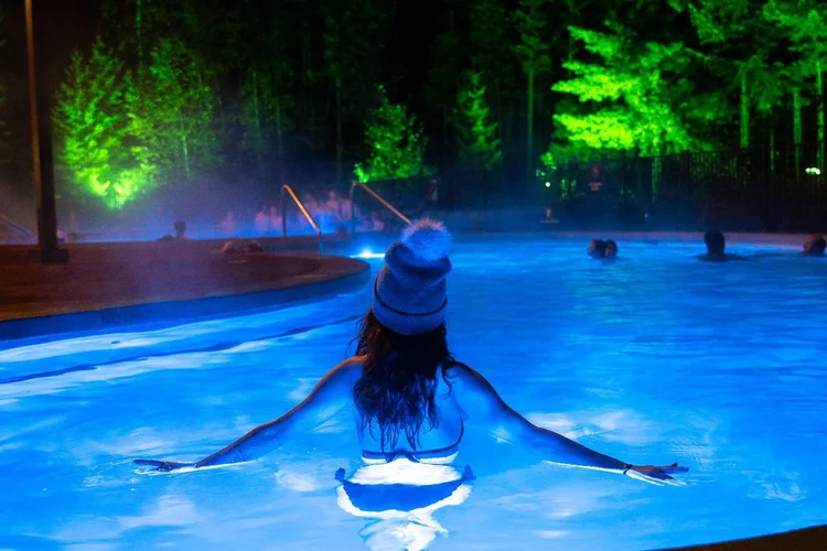 A guest in a grey toque leans on the edge of a blue lit hot pool as steam rises at night
