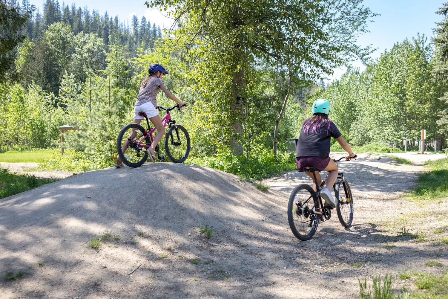 Two kids in helmets riding bikes over the dirt rollers of the Crazy Creek bike track on a sunny day