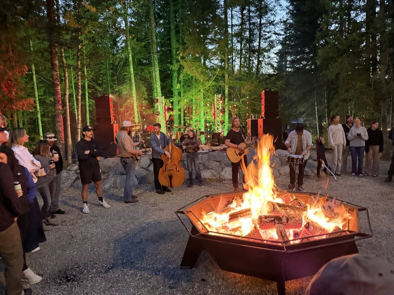 A crowd gathered around the large group fire pit at dusk while a band plays on the stone stage under the lit trees