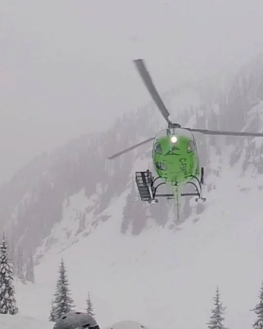 A green helicopter landing in falling snow above skiers in helmets
