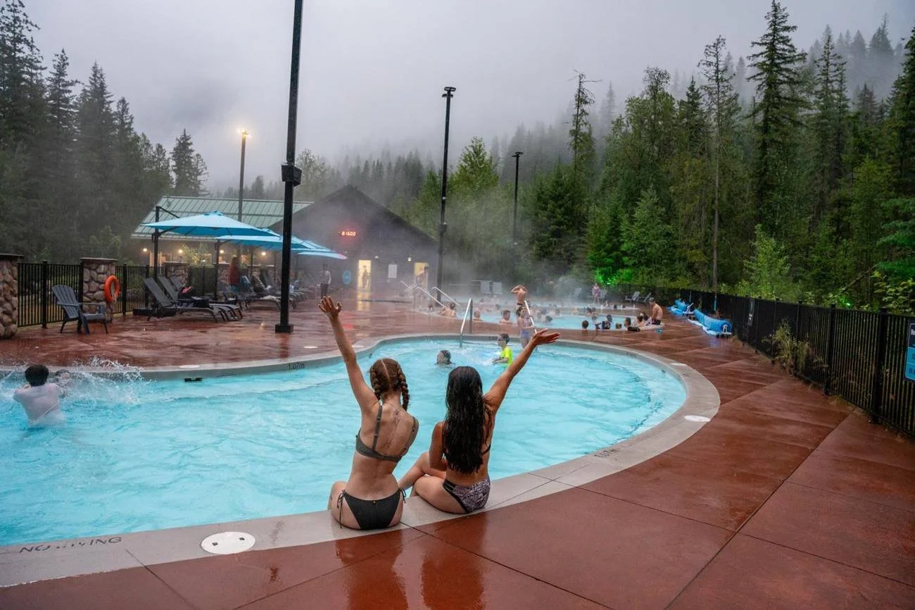 Two friends sitting on the edge of the steaming hot pool with their arms up in the rain, the wet deck shining and cloud on the trees