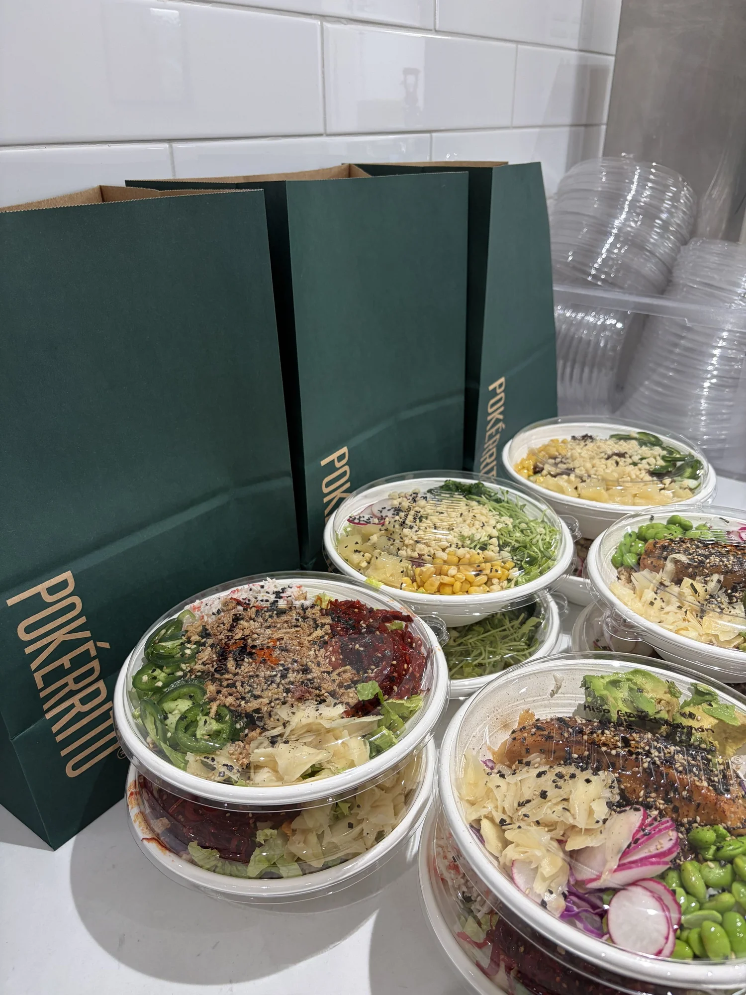 Ten Pokerrito poke bowls with clear lids lined up beside dark green Pokerrito bags, ready for the Compass Group Canada team to pick up
